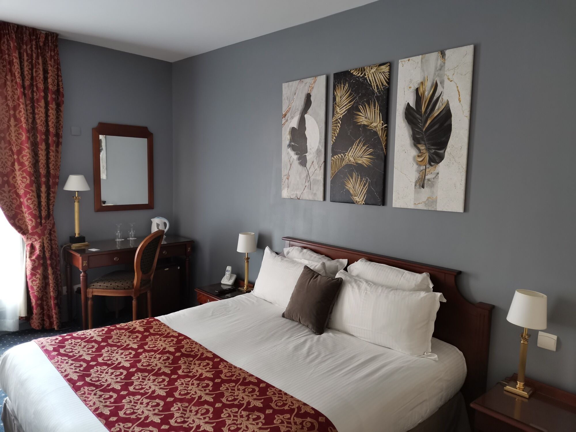 A hotel room with a large bed, a desk with a lamp, a telephone, and a mirror.