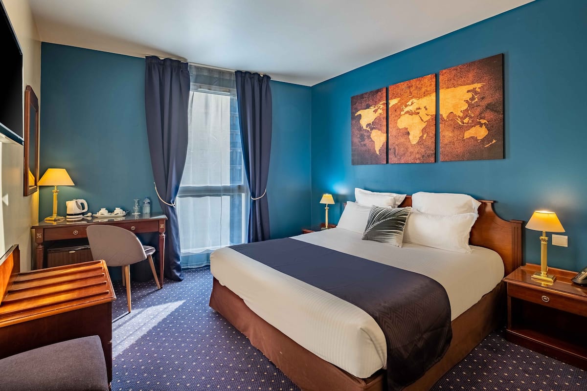A hotel room with a large bed, two bedside tables with lamps, a desk with a chair, a large window with curtains, and a world map artwork on the wall.