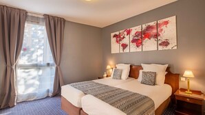 A double bed with a patterned bedspread, a nightstand with a lamp, a wall art piece, and a window with curtains. - Amiral Hôtel (Paris)