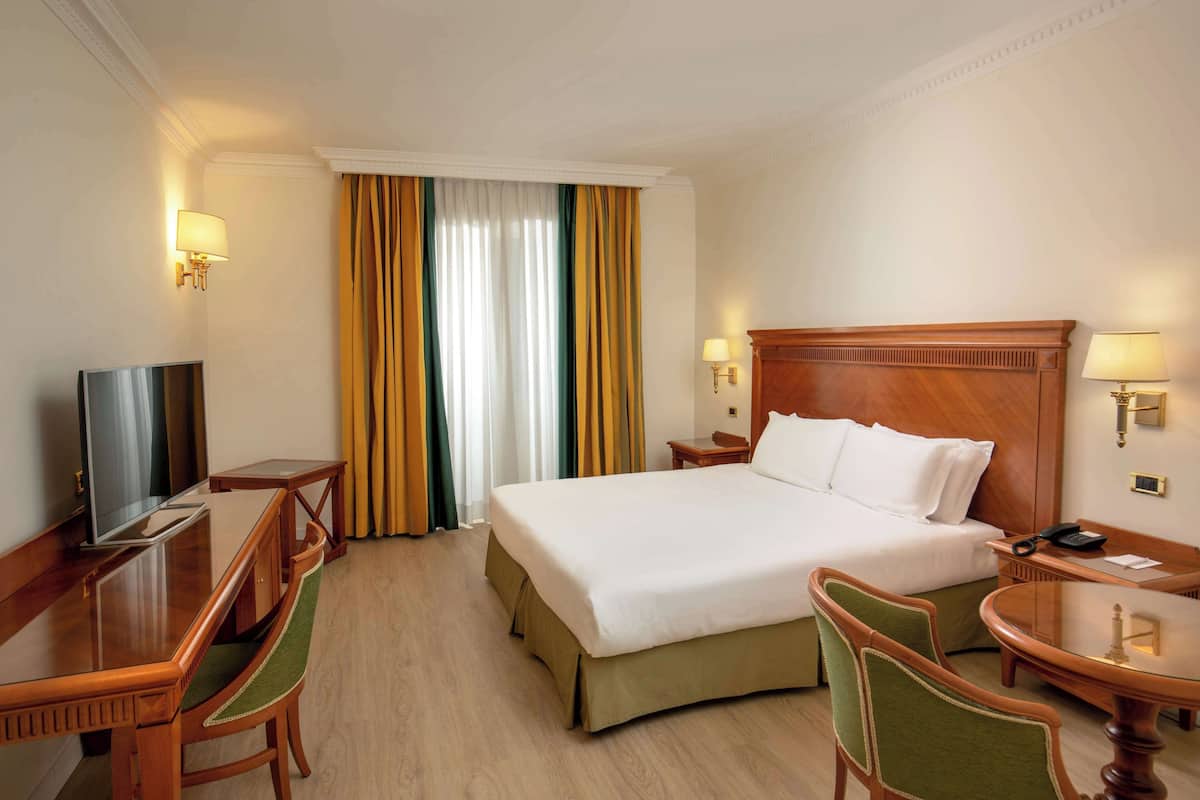 A hotel room with a bed, a desk, a chair, a TV, and a lamp.