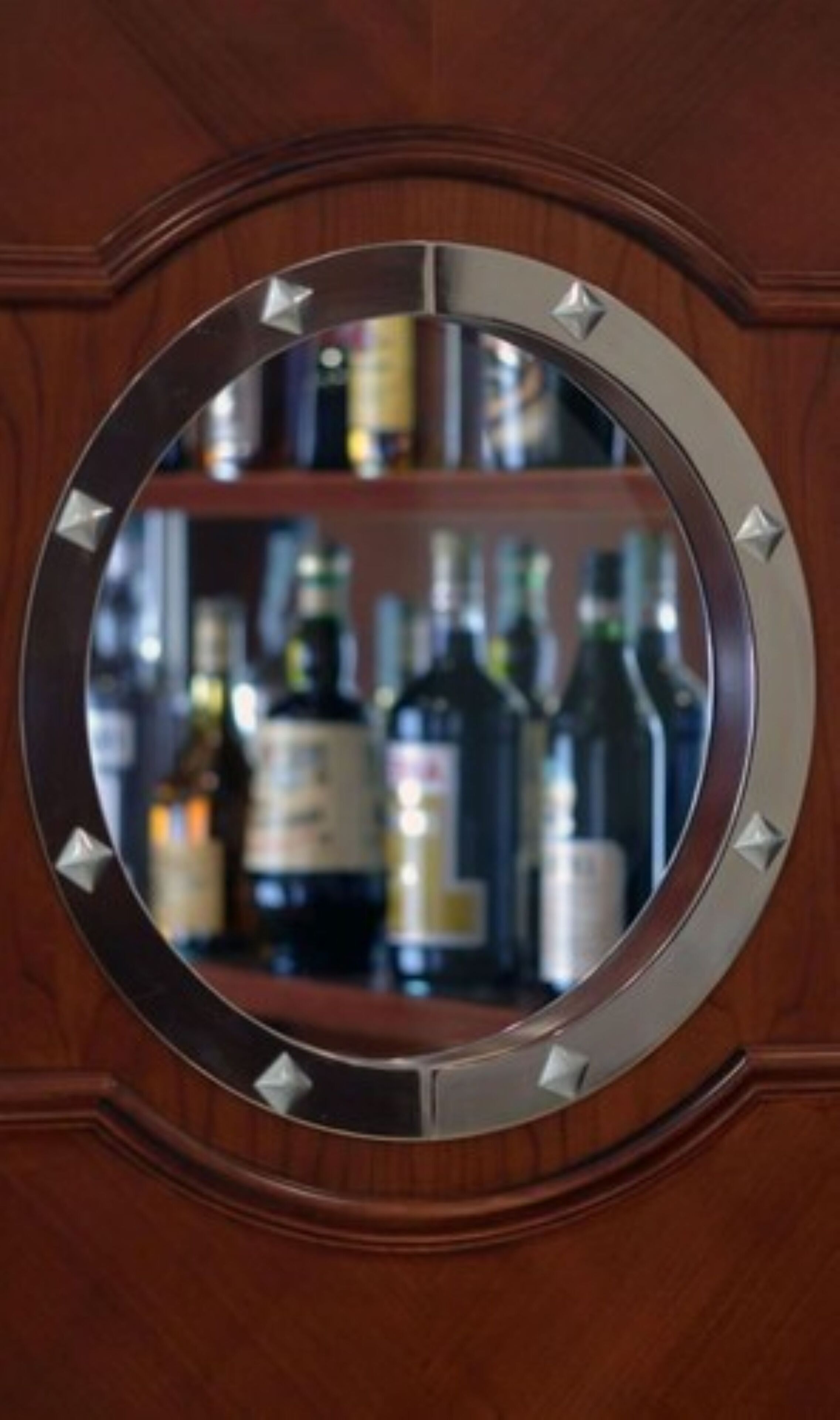 Um espelho circular com moldura metálica, refletindo um interior desfocado com prateleiras de garrafas.