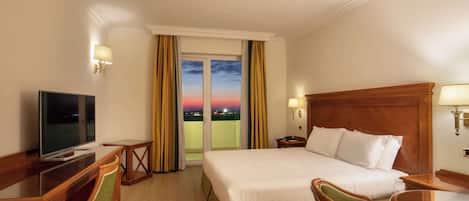 Habitación de hotel con cama, escritorio con televisor y vista a la ciudad al atardecer.