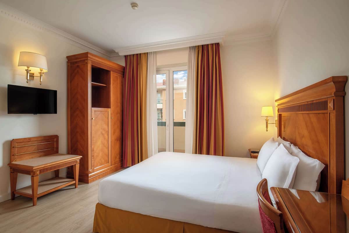 A hotel room with a bed, a wooden nightstand, a television mounted on the wall, and a large window with curtains.