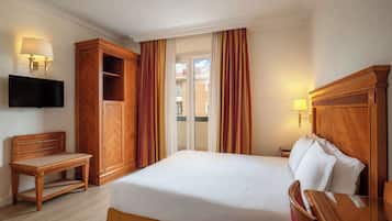A hotel room with a bed, a wooden nightstand, a television mounted on the wall, and a large window with curtains.