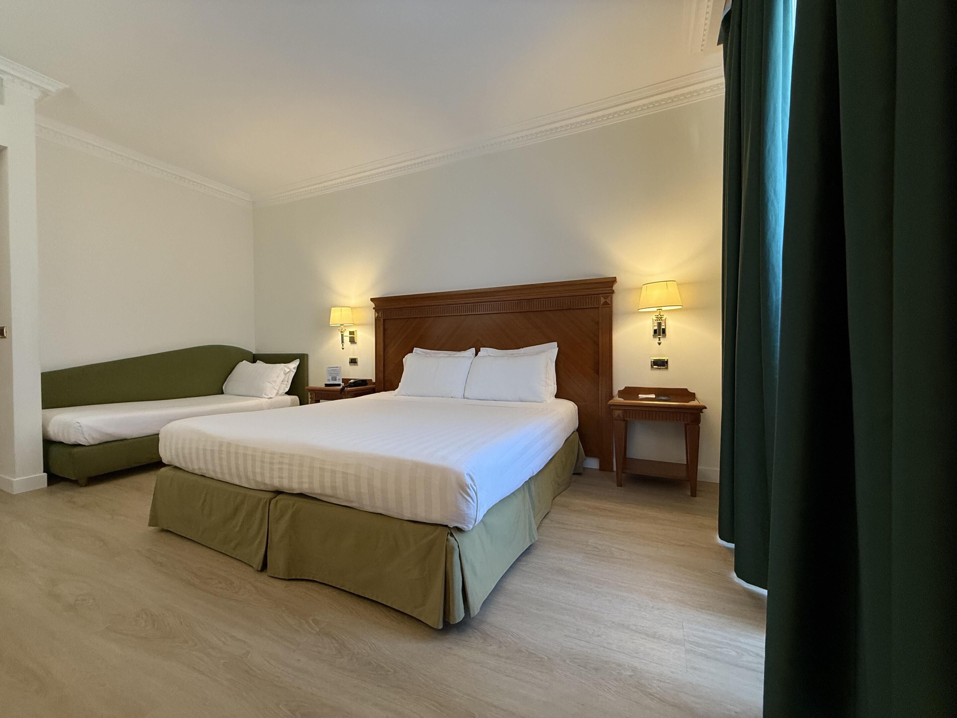 Een hotelkamer met een groot bed, een kleiner bed, twee nachtkastjes en een bijzettafeltje.