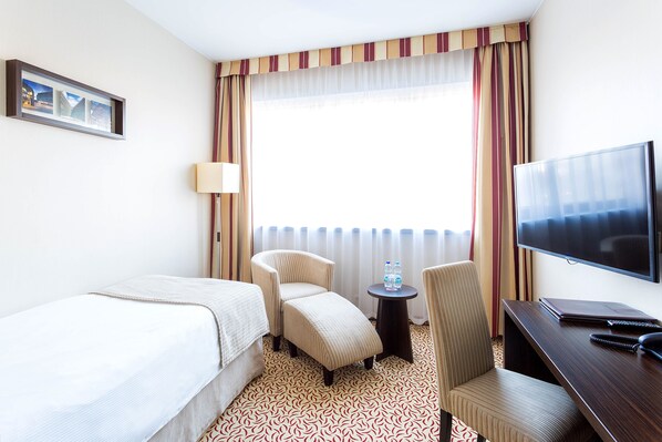 Habitación de hotel con cama, dos sillones, escritorio con televisor y ventana con cortinas.