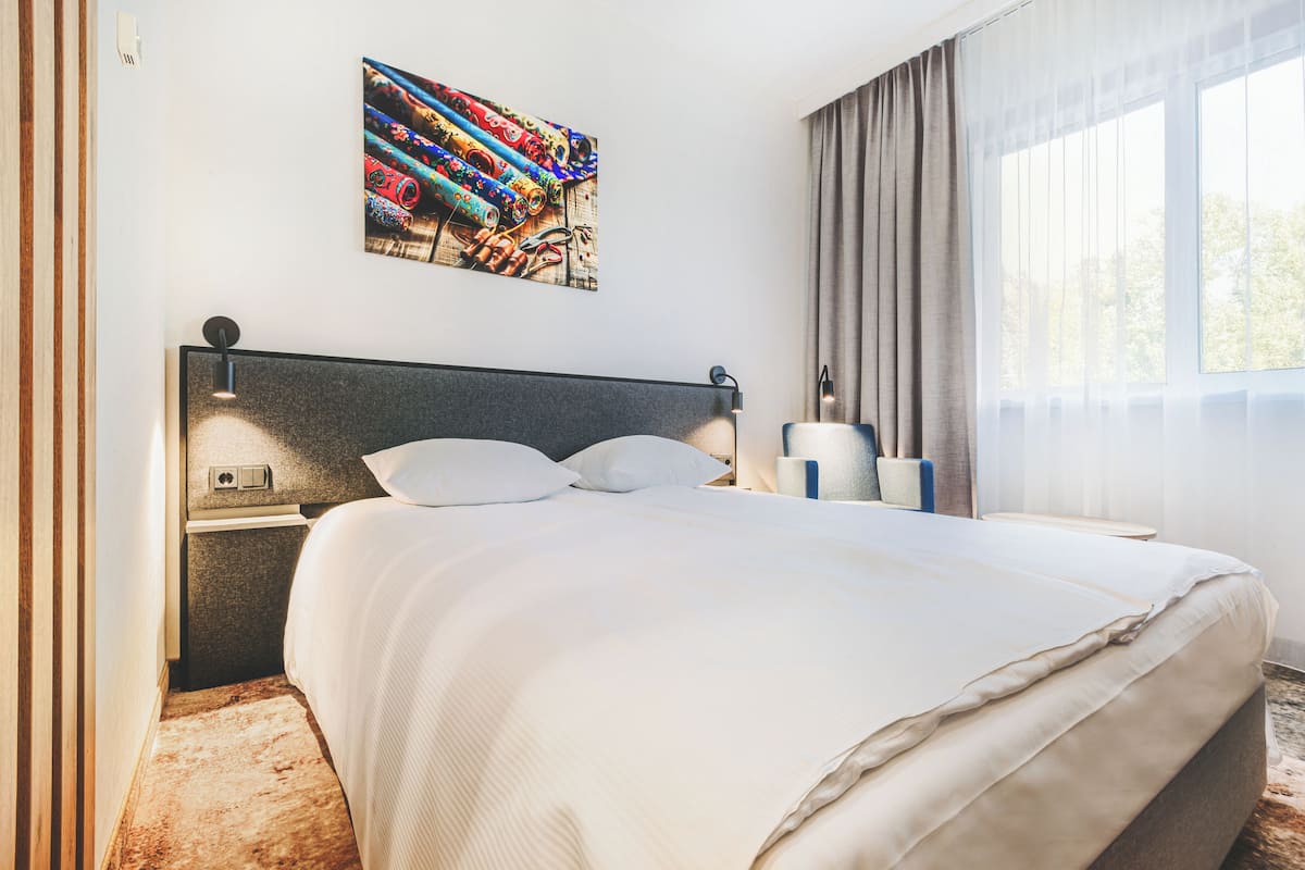 A hotel room with a large bed, a nightstand, a lamp, a painting on the wall, and a window with curtains.
