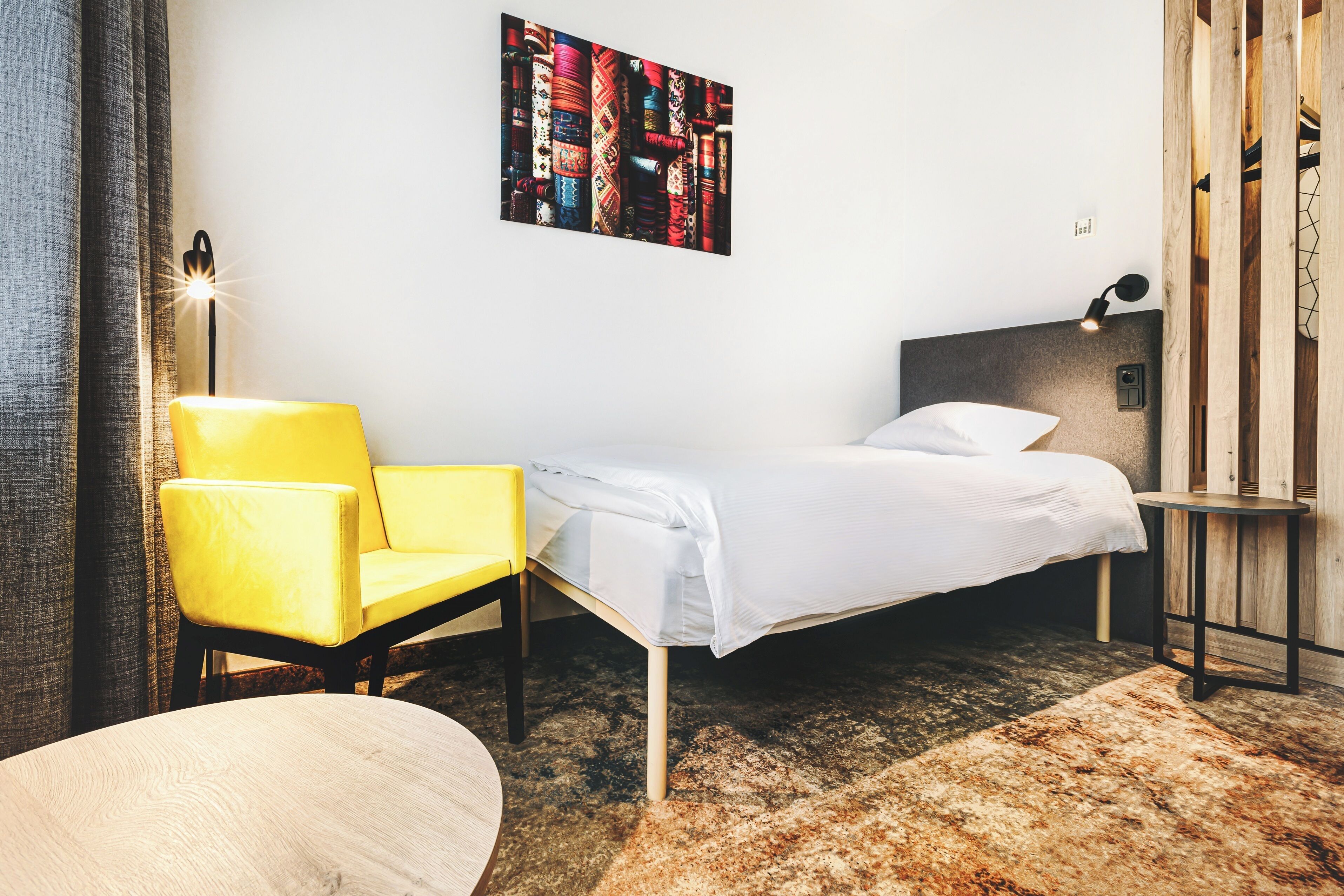 A bedroom with a single bed, a yellow chair, a round table, and a wall-mounted lamp.