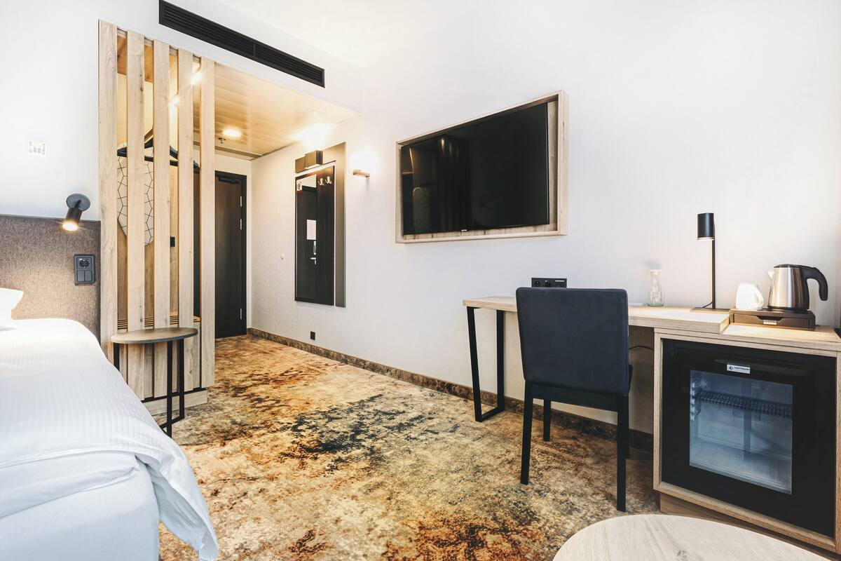 A hotel room with a bed, a desk with a chair, a refrigerator, and a large window.