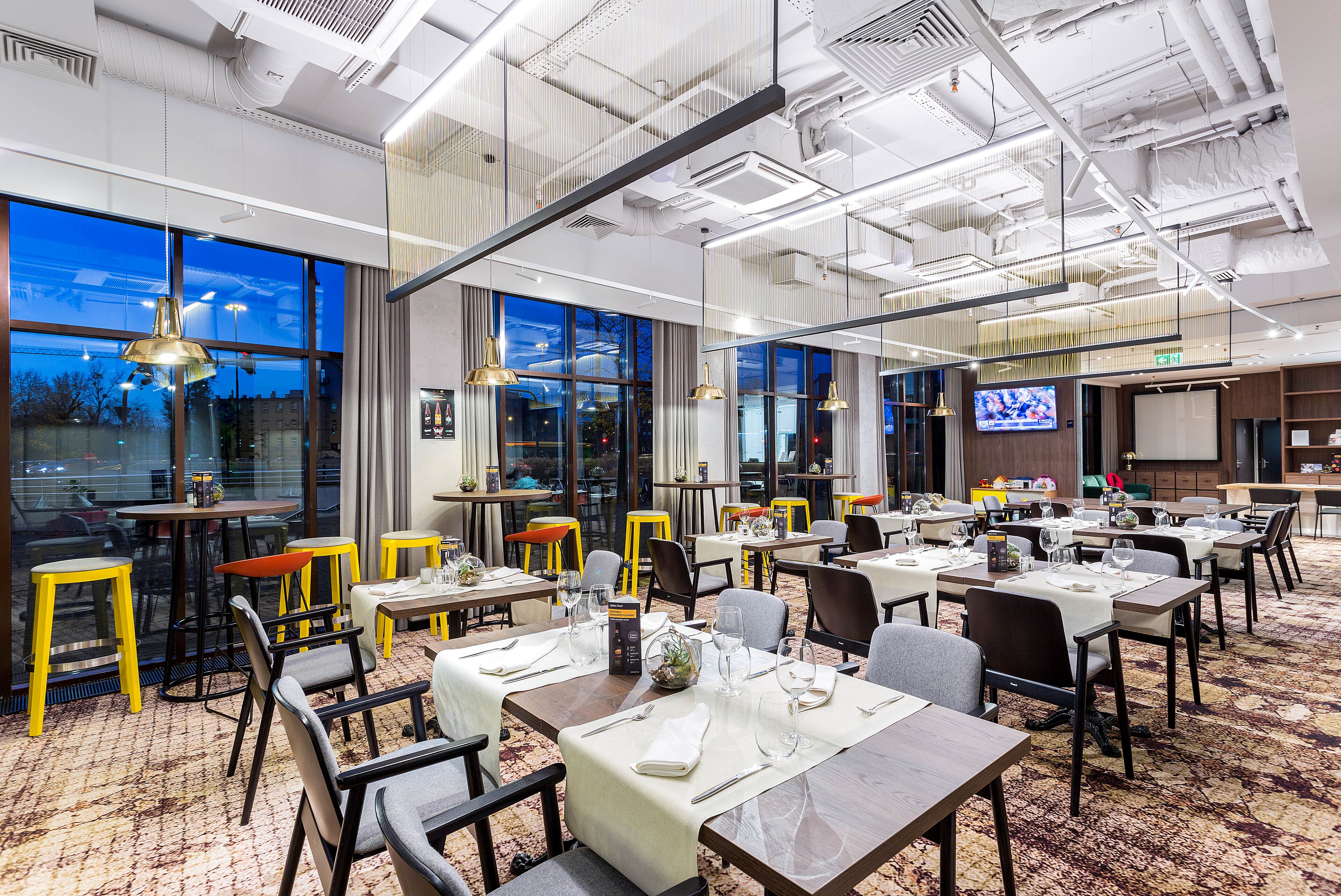A modern dining area with tables set for service, large windows, and a television screen.