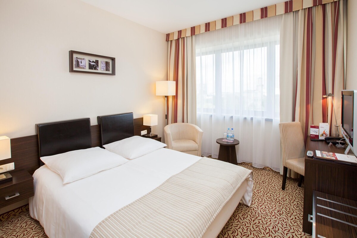 Habitación de hotel con una cama grande, dos mesitas de noche, una silla, un escritorio con computadora y una ventana con cortinas.