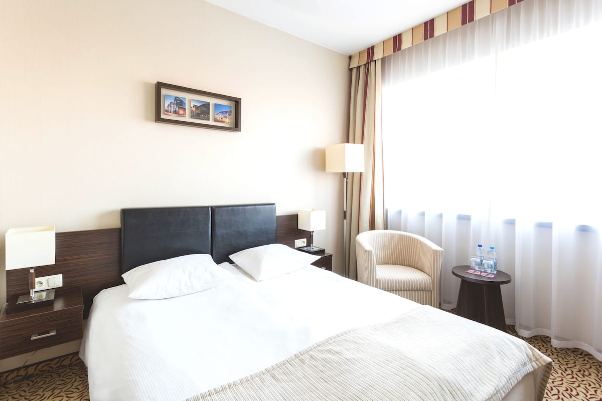 A hotel room with a large bed, a chair, a small table with water bottles, and a picture on the wall.