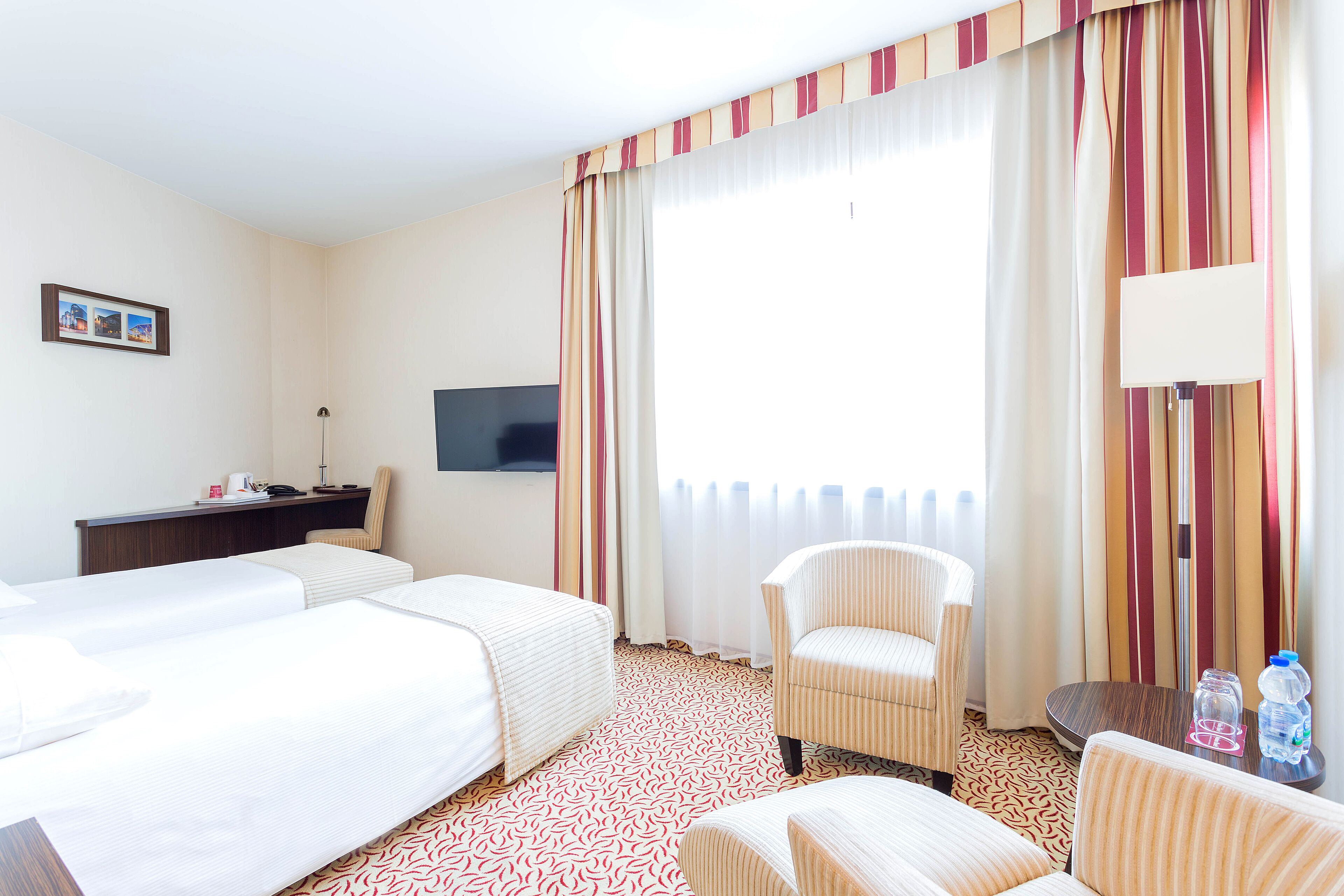 A hotel room with two beds, a desk, a chair, a TV, and a window with curtains.