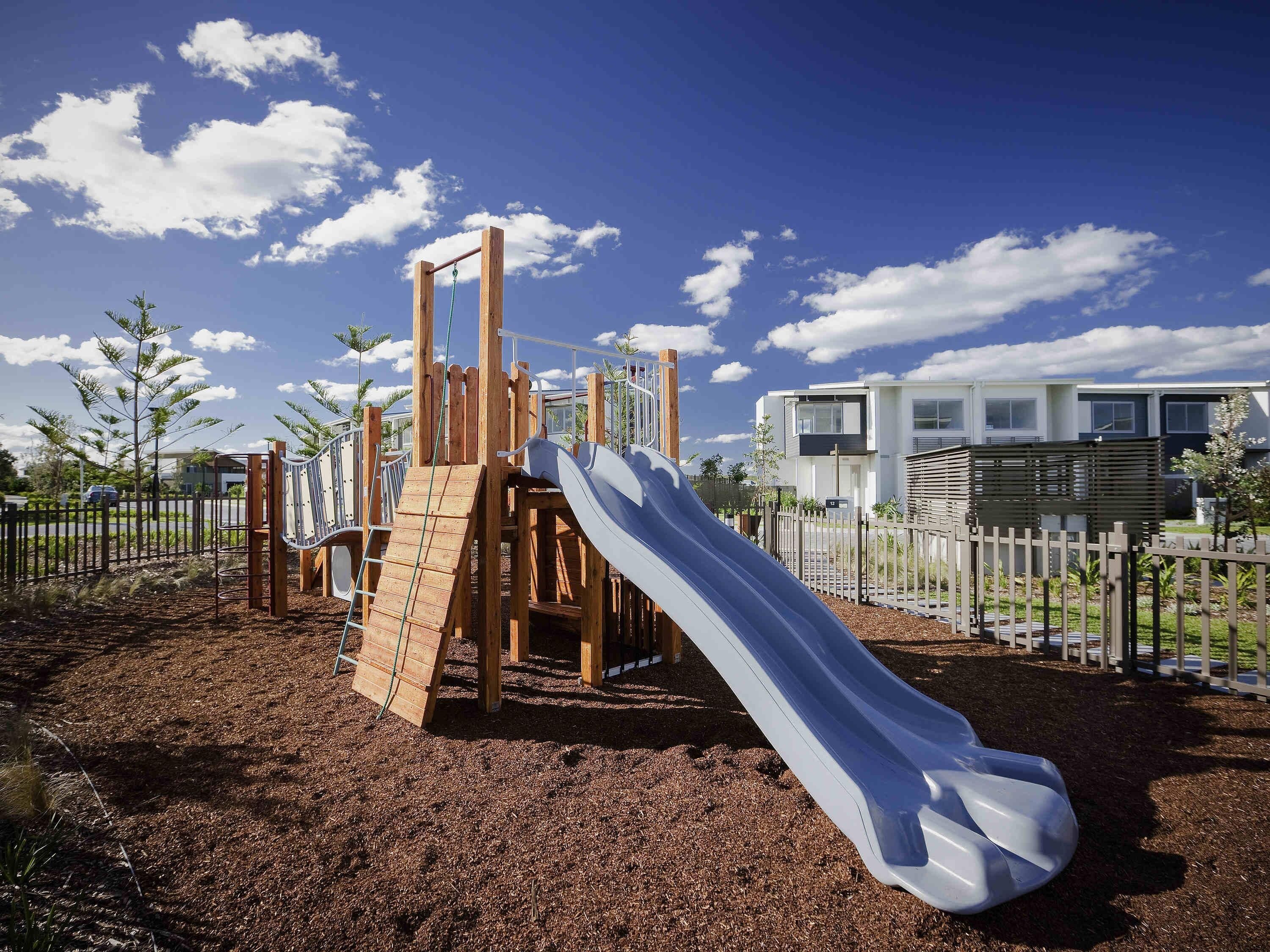 A playground with a slide and climbing structures, surrounded by a fence and trees.