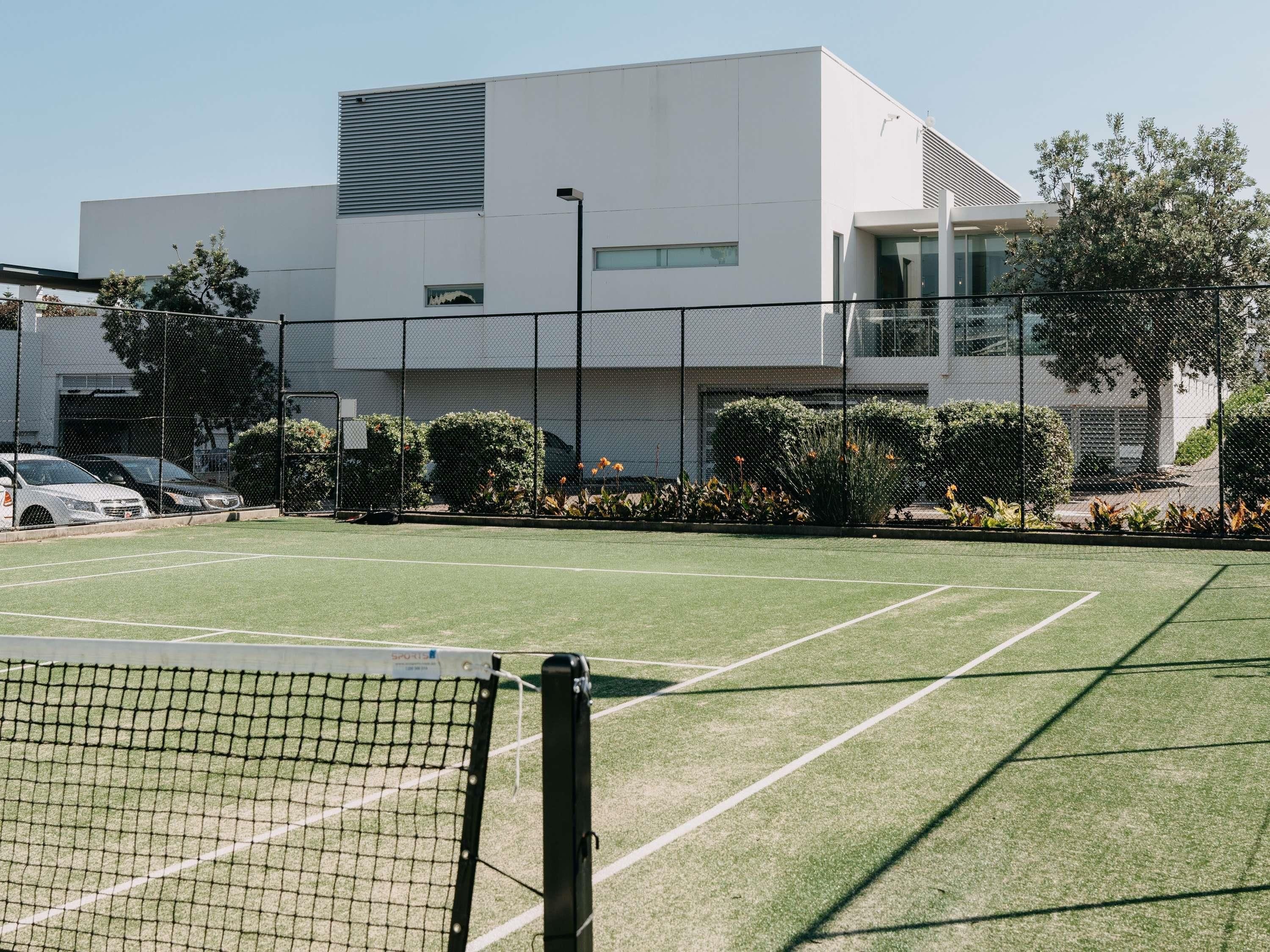 Una cancha de tenis con red, rodeada por una valla y árboles.