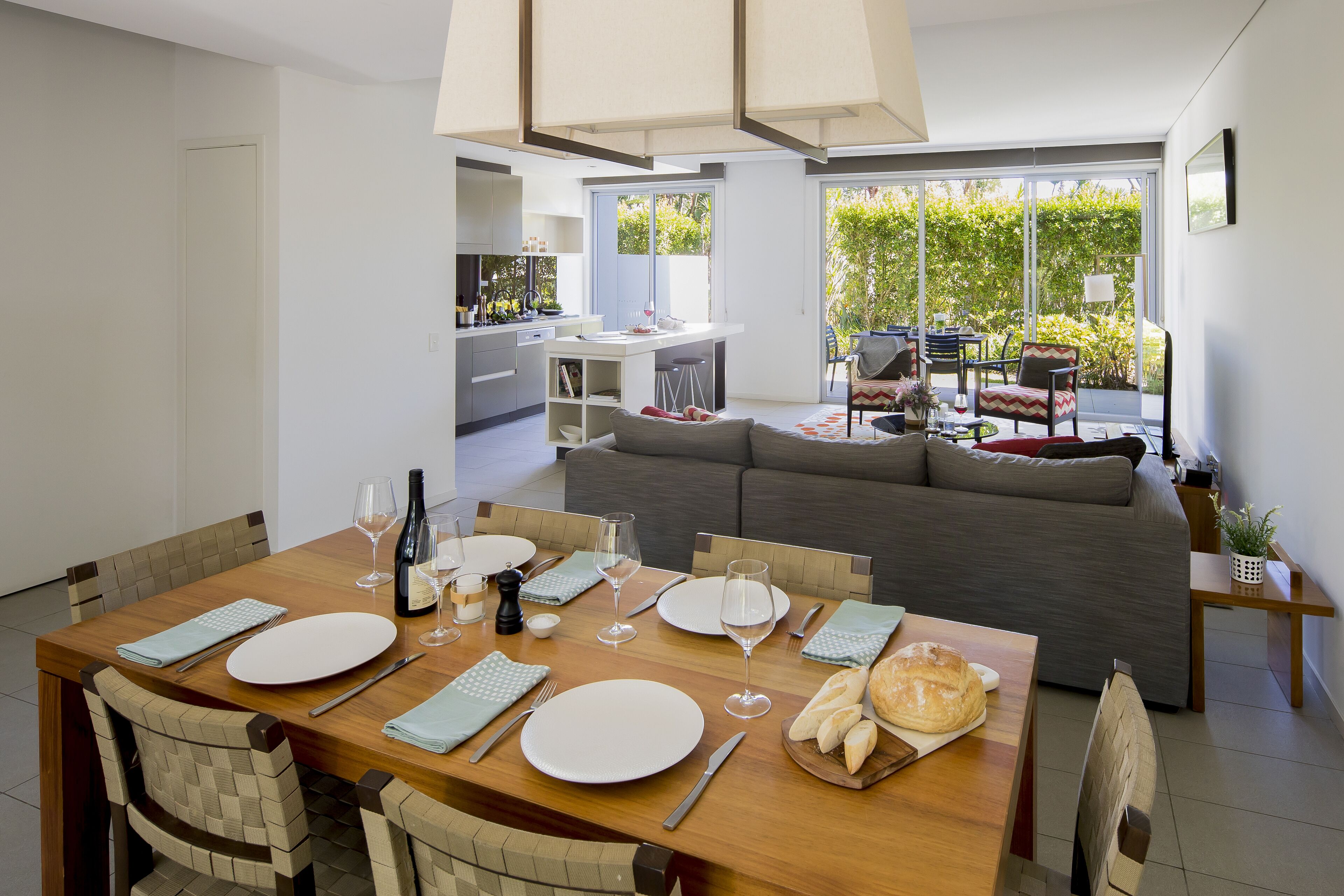 A modern dining area with a wooden table set for four, a grey sofa, and a kitchen in the background.