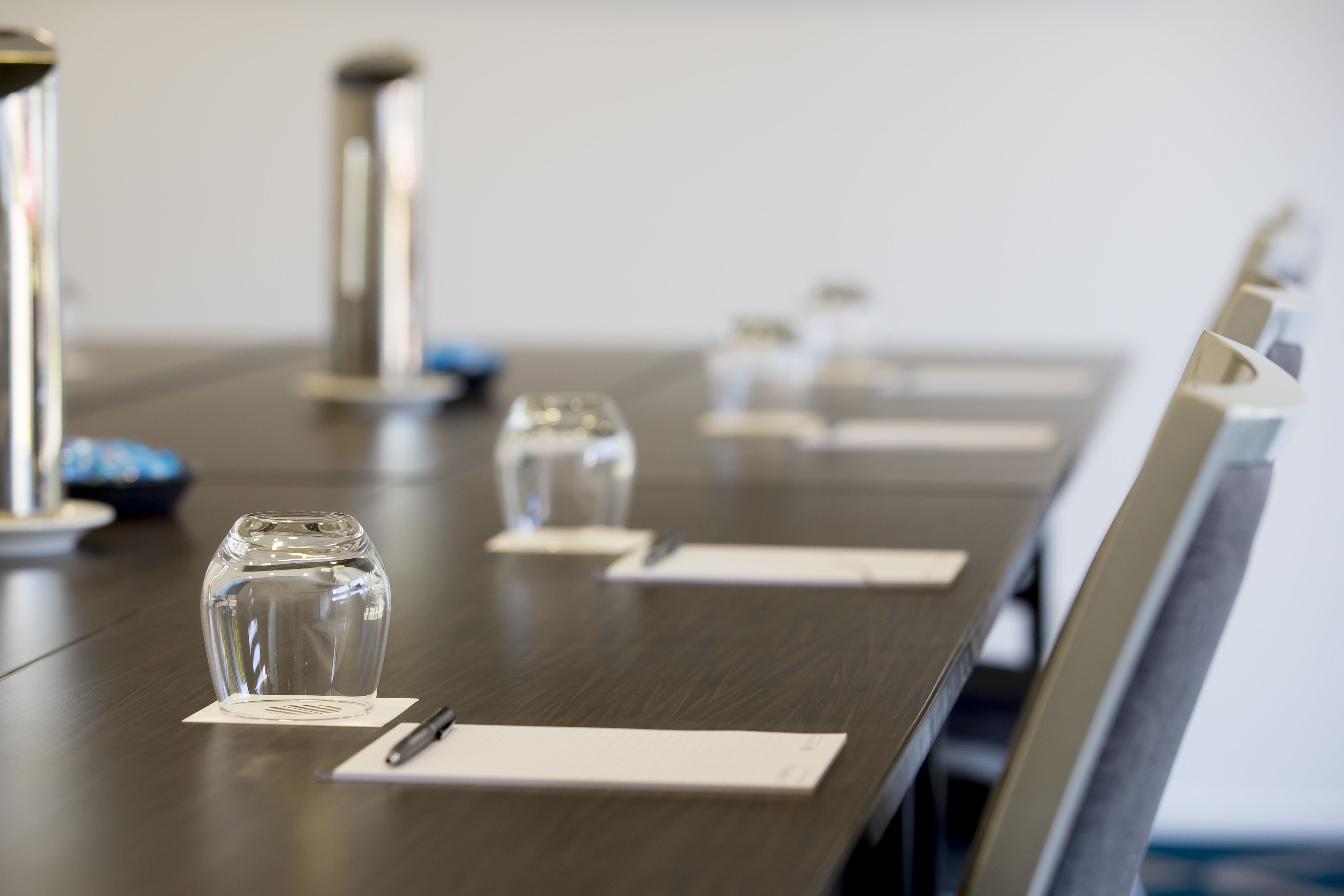 Mesa de conferência com copos de vidro transparentes e canetas, sobre um fundo neutro.