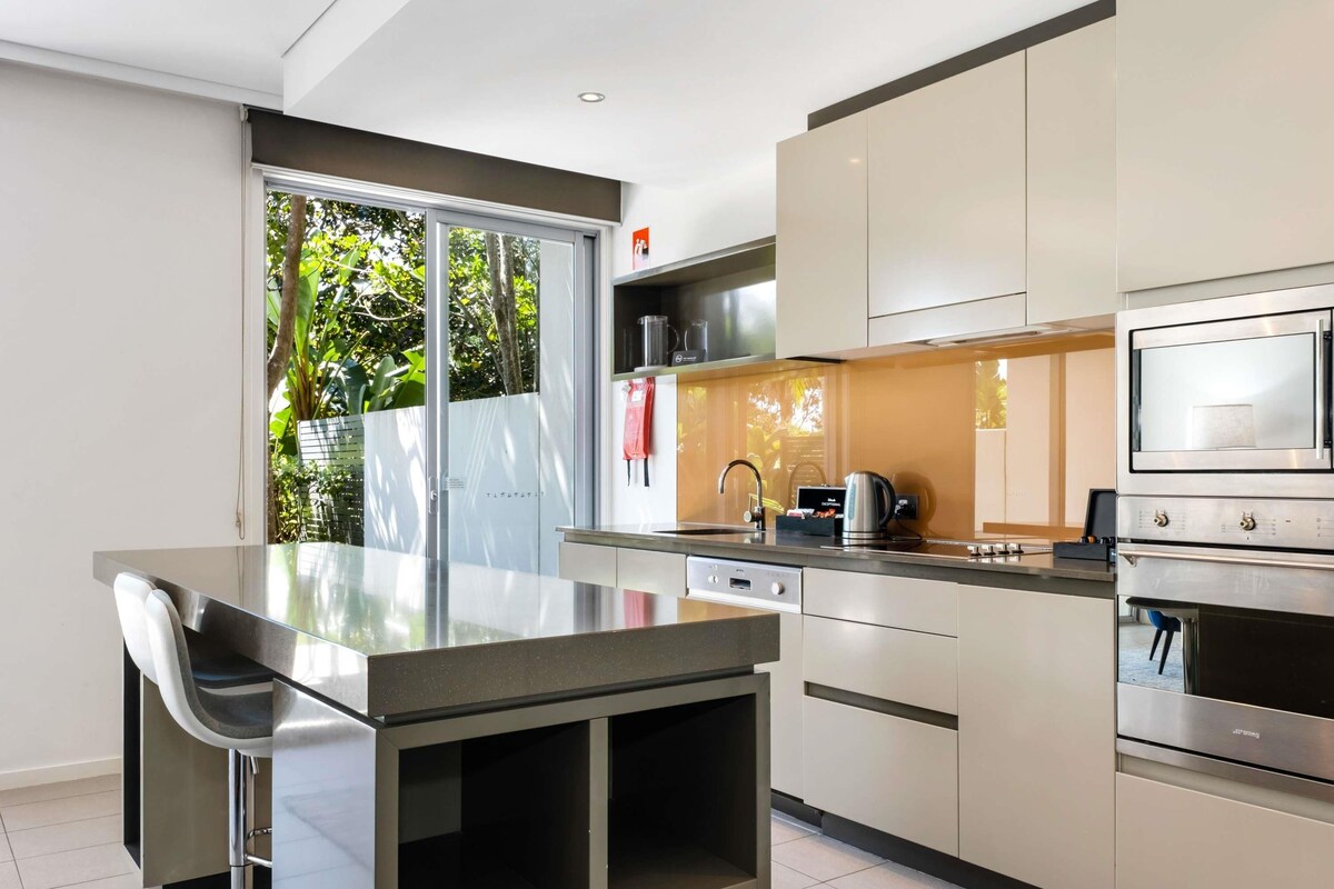 A modern kitchen with a glass dining table, stainless steel appliances, and a view of greenery outside.