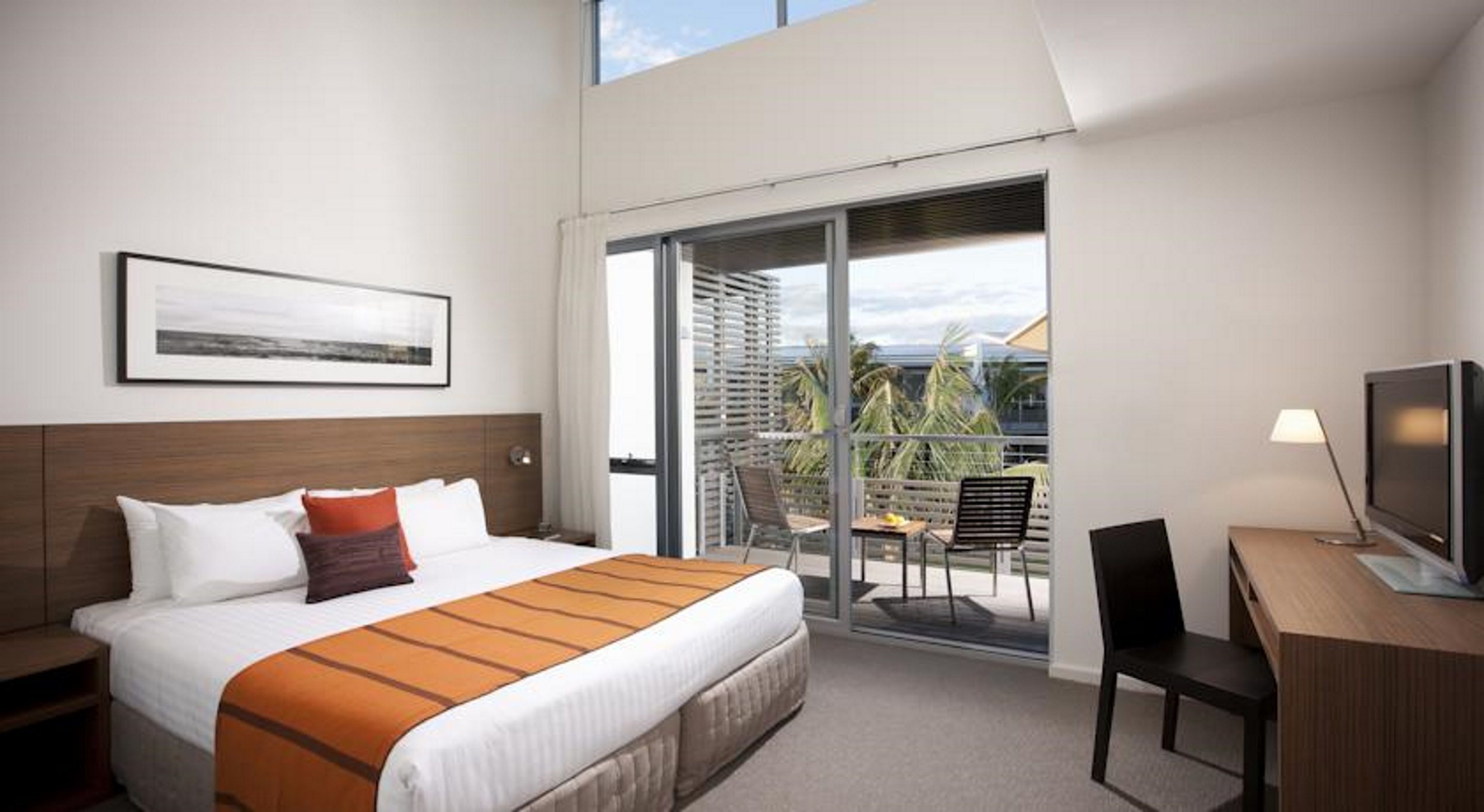 Ein Hotelzimmer mit Bett, Schreibtisch mit Fernseher, Stuhl und Balkon mit Blick auf Palmen.