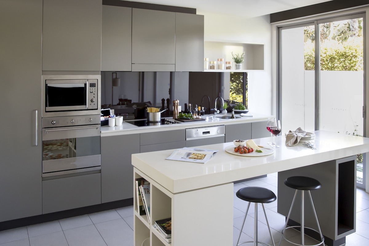 A modern kitchen with built-in appliances, a central island with stools, and a view through a sliding door.