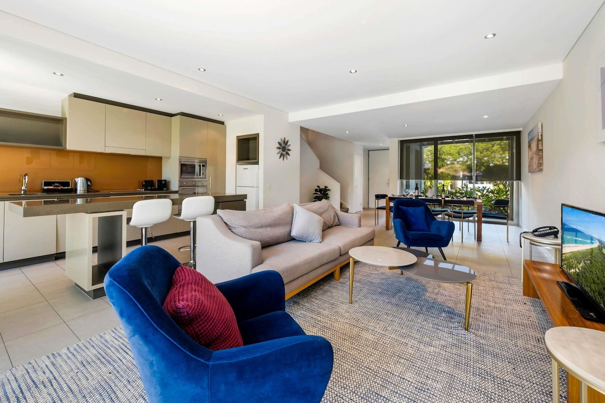 Un salon moderne avec un canapé, un fauteuil bleu et une table basse. Il y a également un coin cuisine équipé d’un four à micro-ondes et d’un évier.