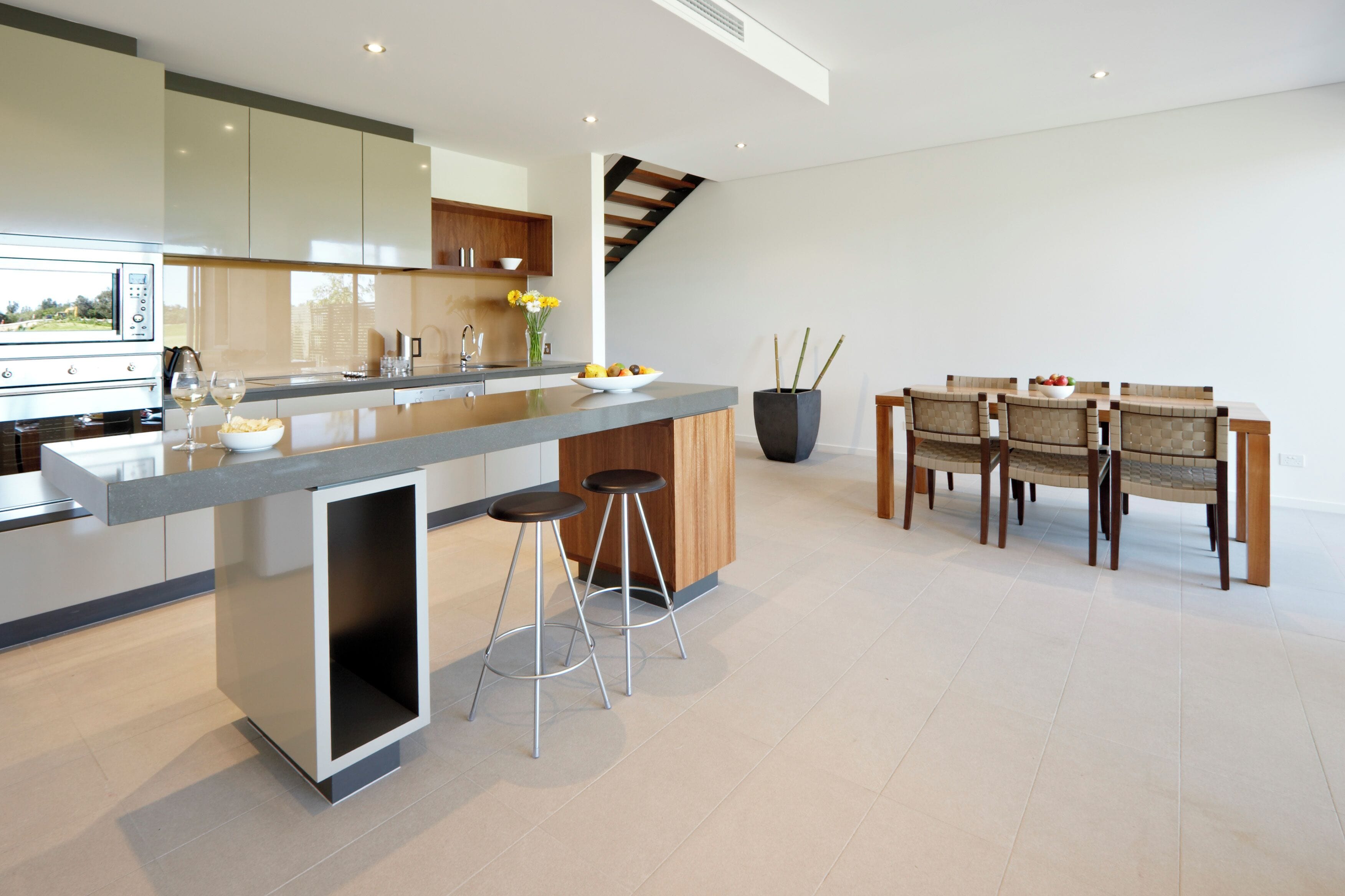 A modern kitchen with a central island, bar stools, a dining area with wooden chairs, and a large potted plant.