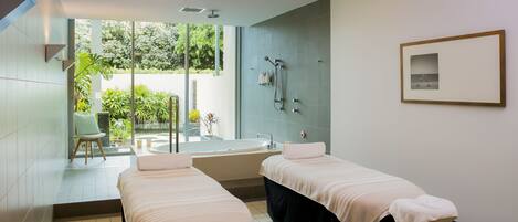A modern spa room with two massage tables, a large bathtub, and a view of greenery outside.