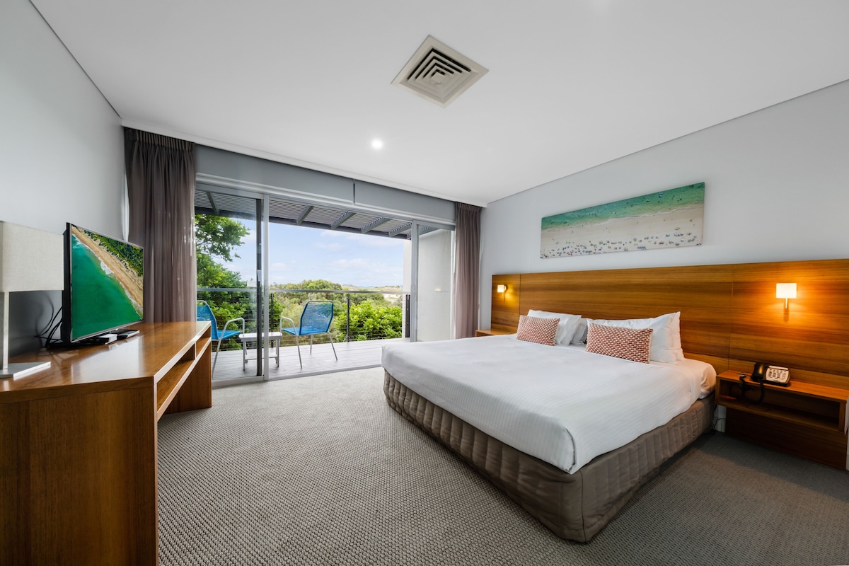 A modern hotel room with a large bed, a desk with a flat-screen TV, a balcony with a view of greenery, and a painting on the wall.
