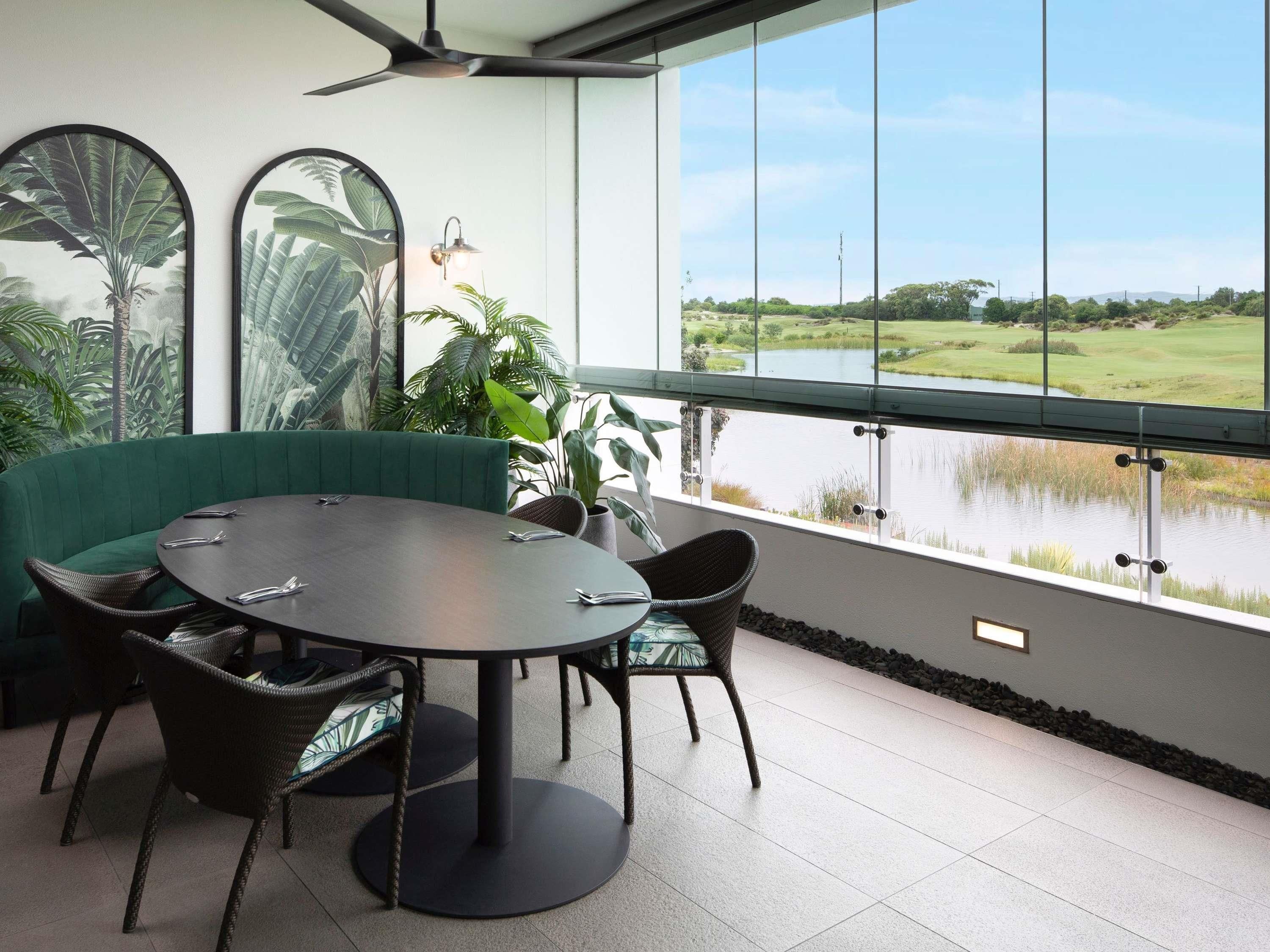 Un espace de restauration moderne avec une table ronde, un canapé vert, des chaises en osier et une vue sur un lac et un terrain de golf.