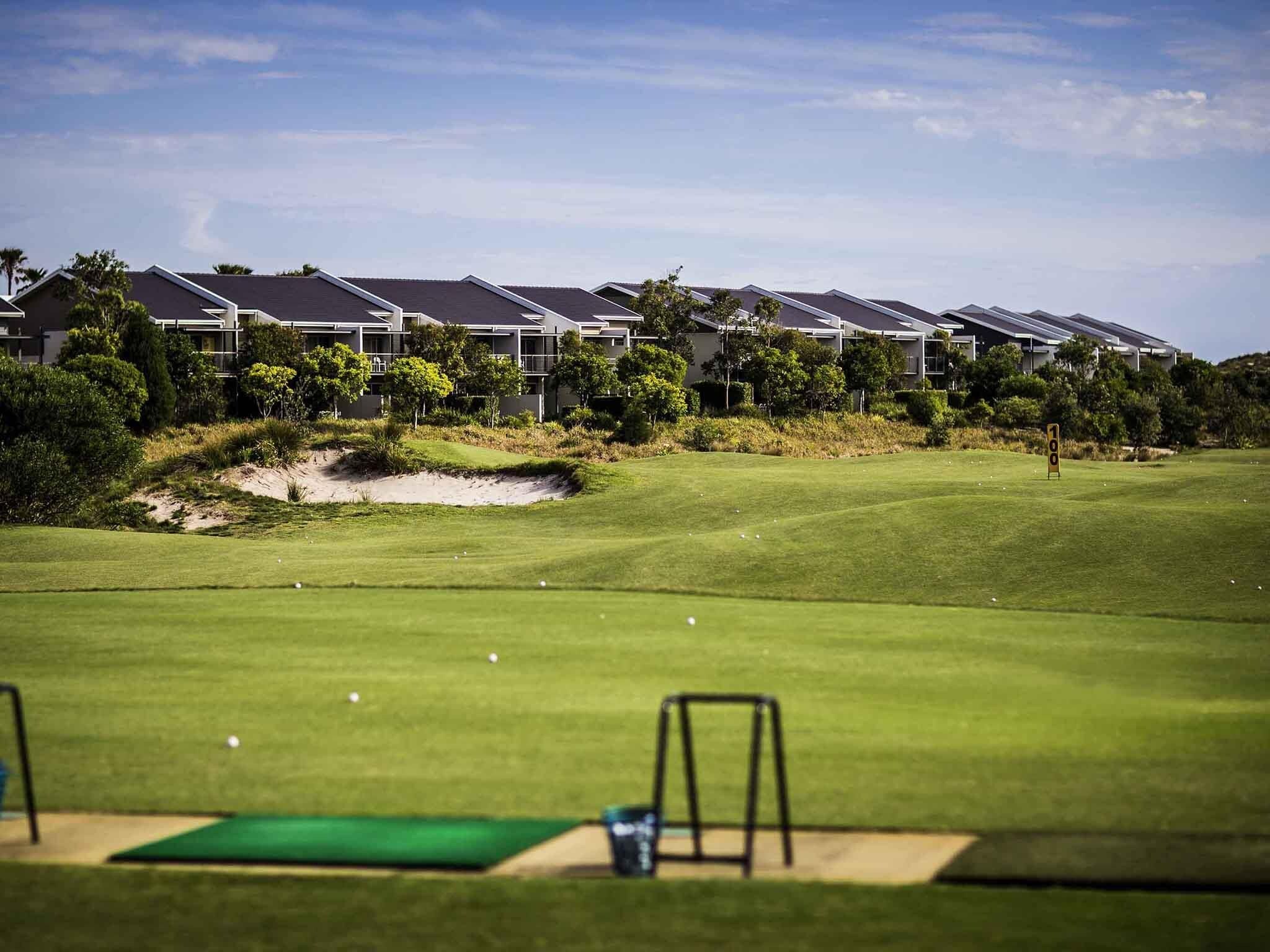Ein Golfplatz mit Putting Green, Sandbunkern und einer Reihe moderner Häuser im Hintergrund.