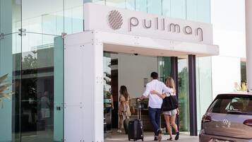 A hotel entrance with a glass facade, a sign reading "Pullman," and people exiting with luggage.
