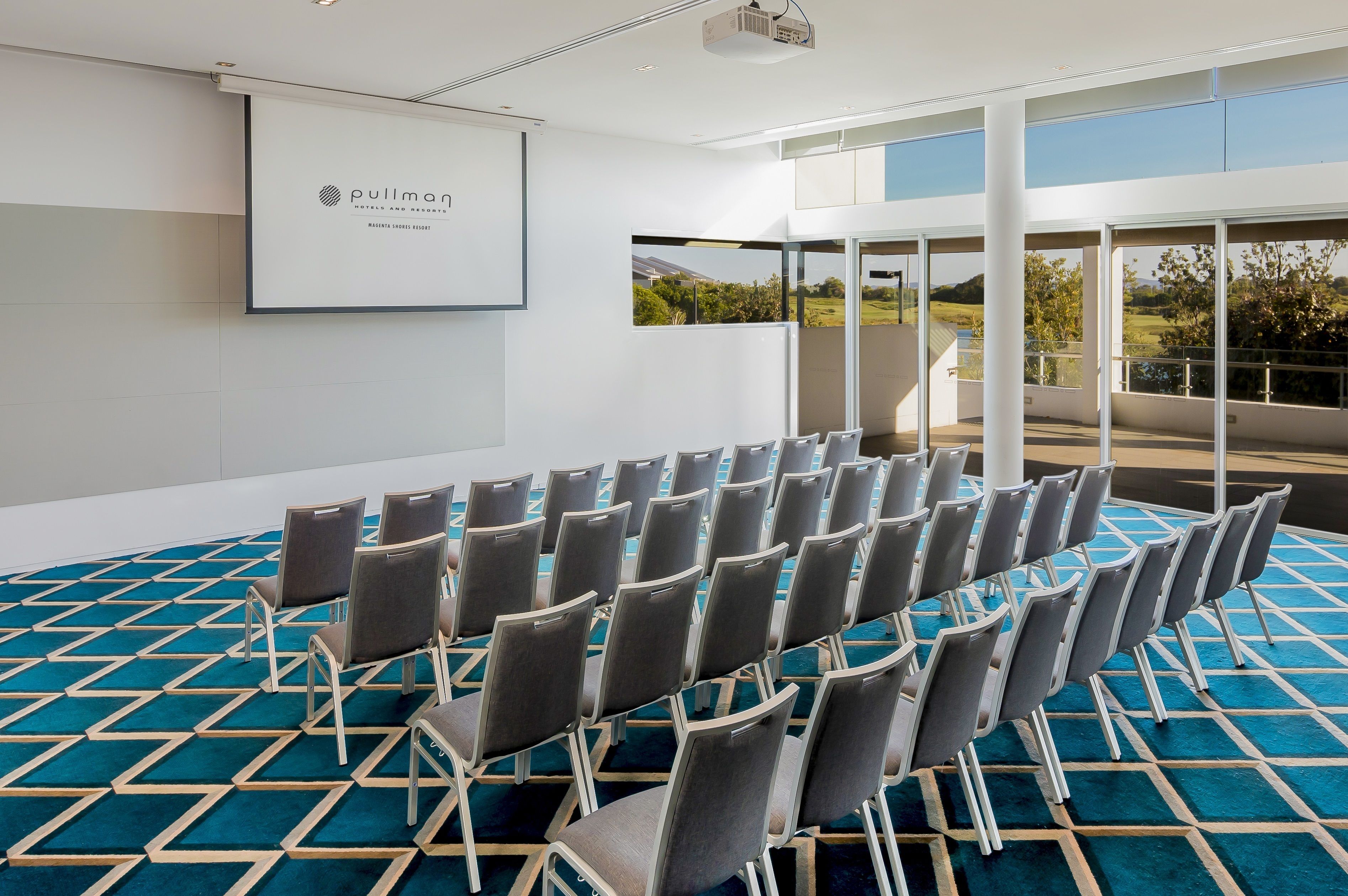 A conference room with rows of chairs, a projector screen, and large windows offering a view of the outdoors.
