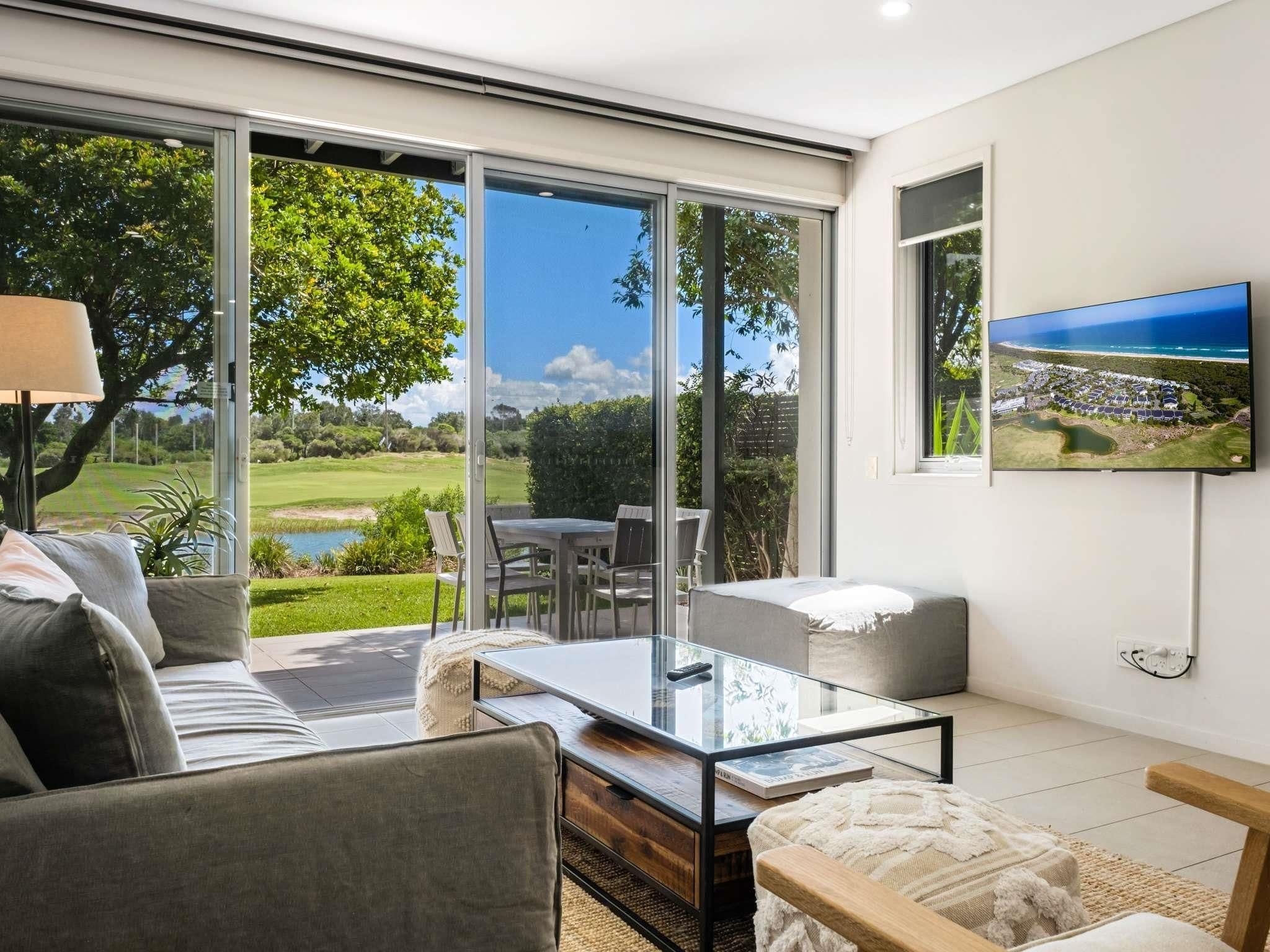 A modern living room with a glass table, comfortable seating, and a large TV displaying a scenic view.