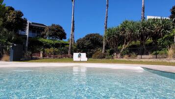 A clear blue pool with a person sitting on a white bench in the center. Tall palm trees and dense foliage surround the pool area.