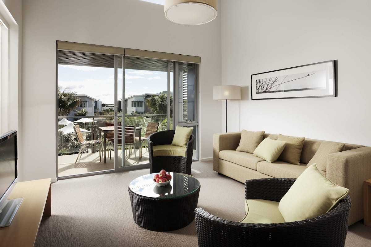Un salon moderne comprenant un canapé, des fauteuils, une table basse en verre, un lampadaire et une grande fenêtre donnant sur l’extérieur.