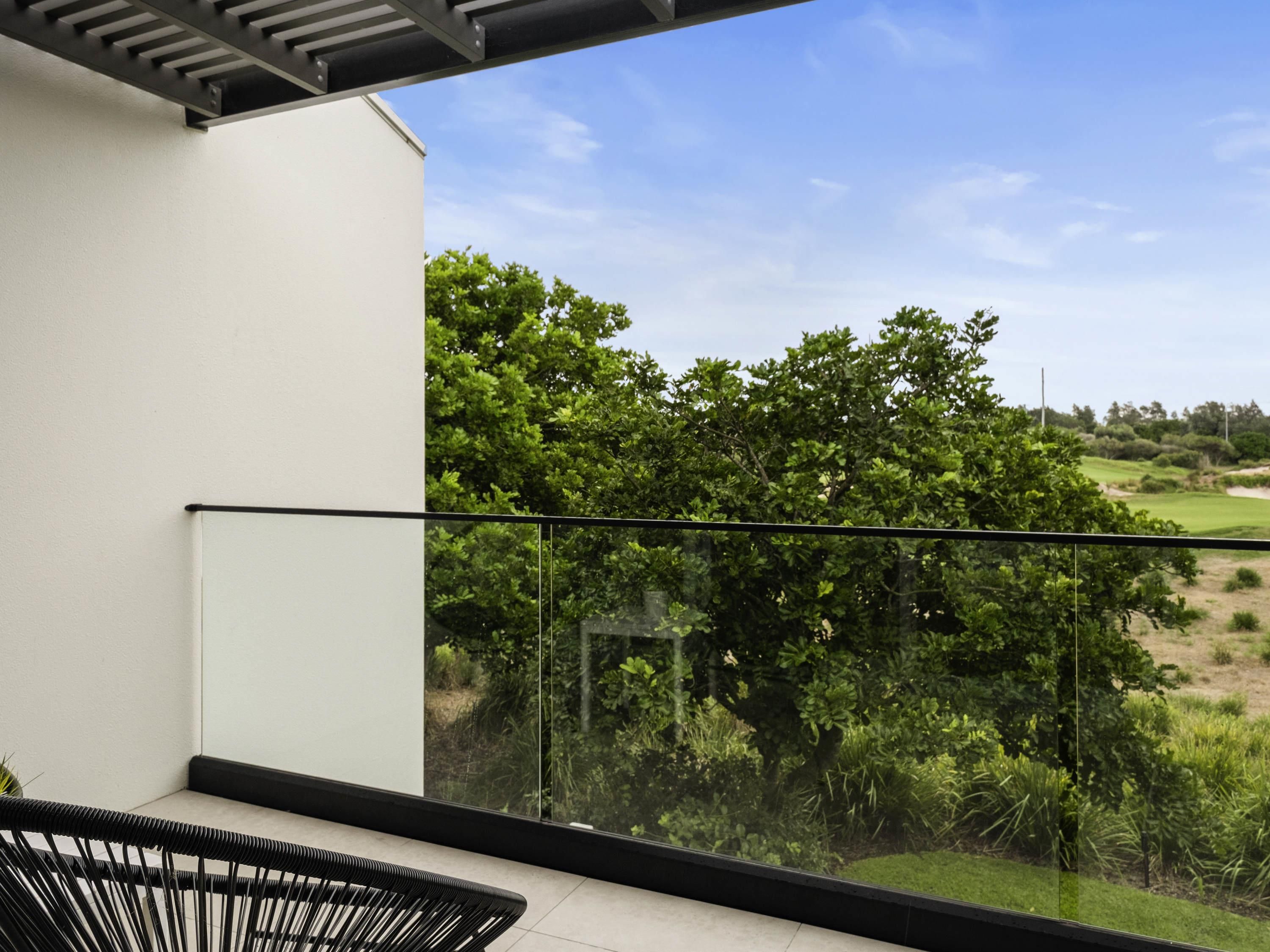 Un balcon moderne doté d’une balustrade en verre, offrant une vue sur un jardin luxuriant et un terrain de golf.