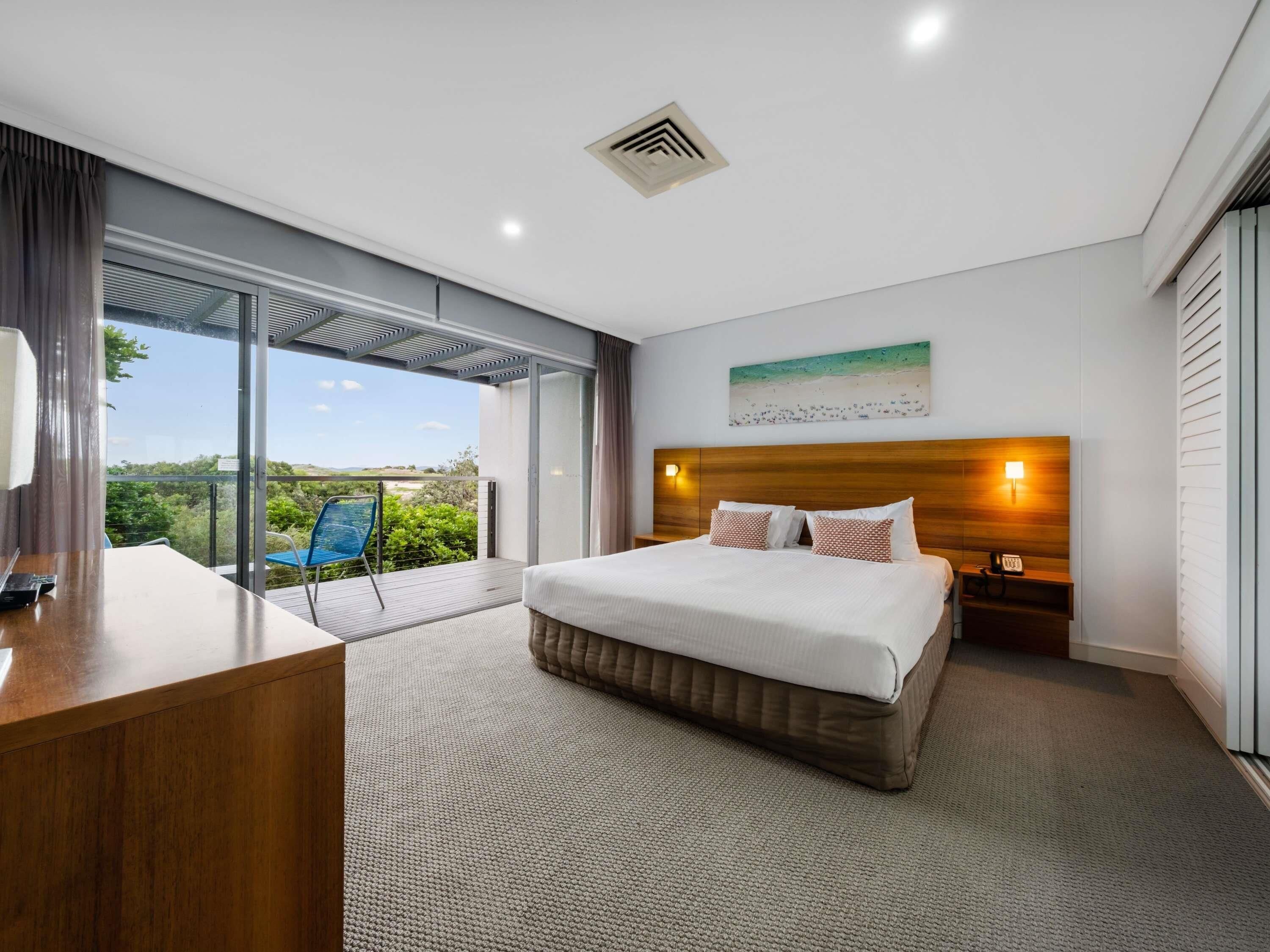 A modern hotel room with a large bed, a desk, and a balcony with a view of the beach.