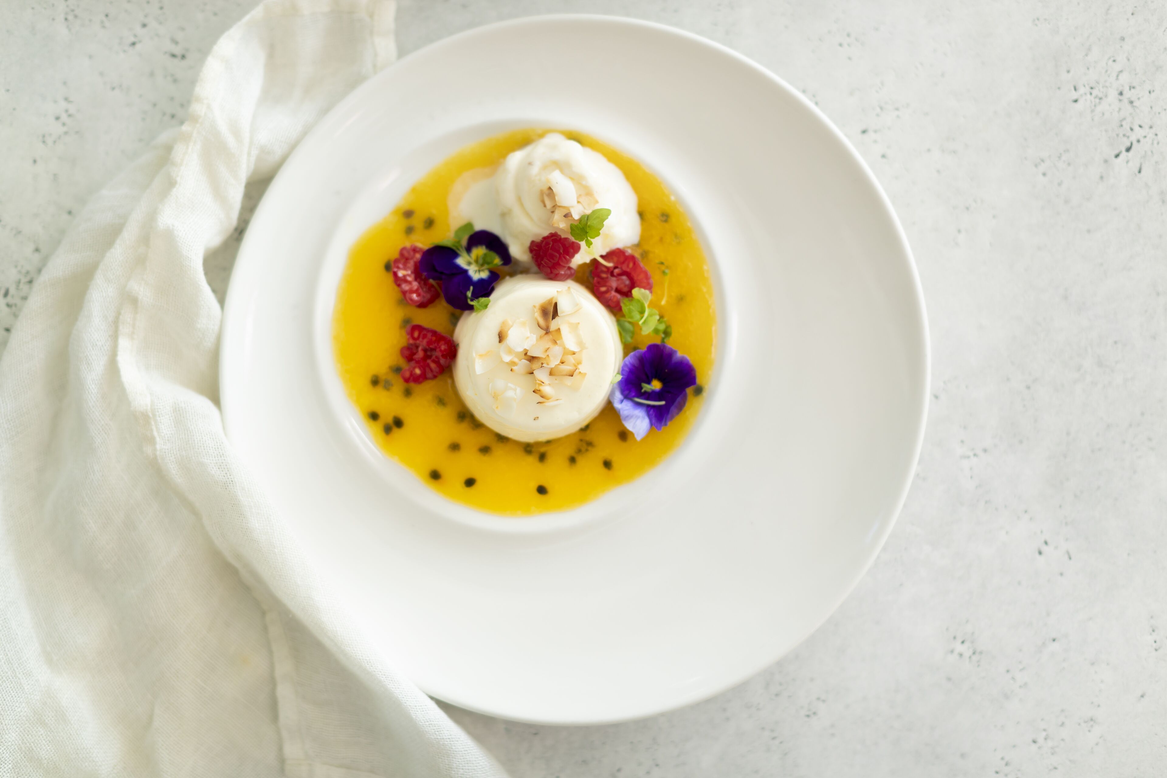 A white plate with two round desserts on a yellow sauce, garnished with raspberries and purple flowers.