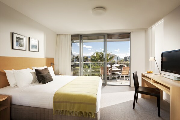 Habitación de hotel con cama, escritorio, silla, televisor y balcón con vista a palmeras y edificios.
