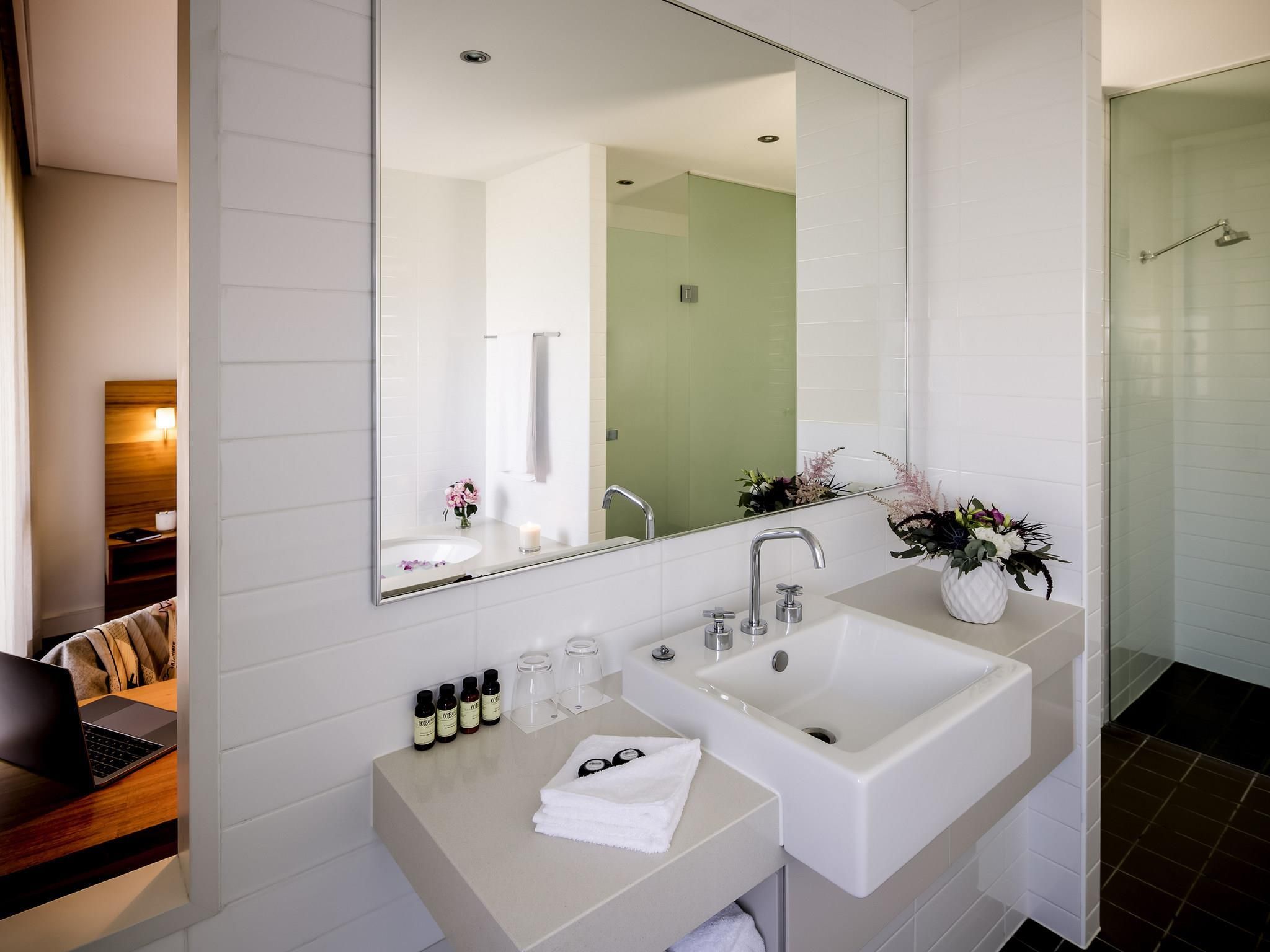 A modern bathroom with a large mirror, two sinks, and a shower. There are toiletries on the counter and a towel rack above the sink.
