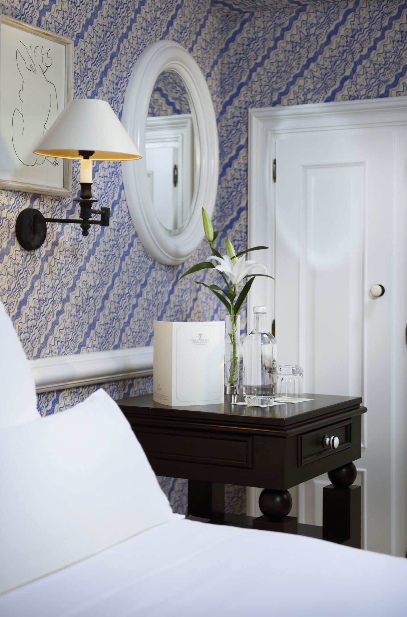 Una habitación de hotel con una cama, una mesita de noche con una lámpara, un jarrón con flores, un espejo y una puerta.