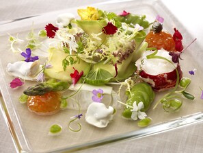 A square glass plate with a variety of food items, including vegetables, flowers, and possibly some meat, arranged artistically.