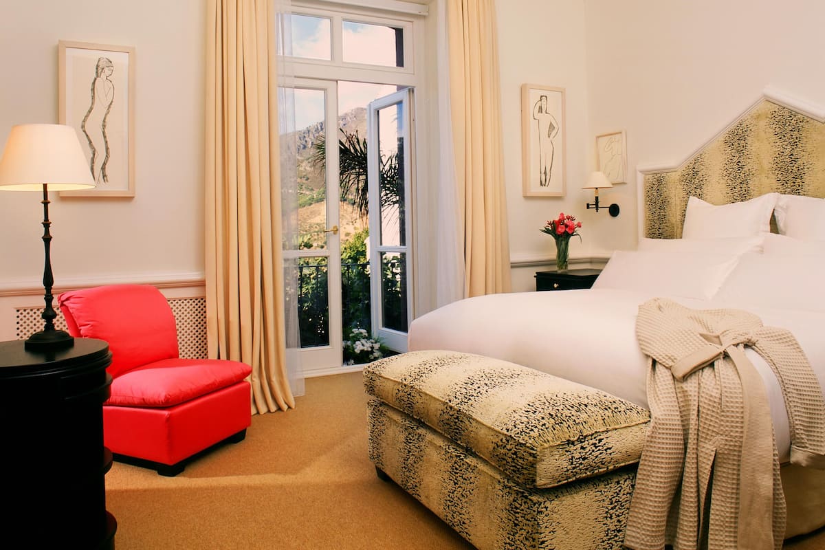 A hotel room with a large bed, a red chair, a small table with a lamp, a window with curtains, and artwork on the walls.