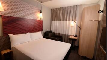 Quarto de hotel com uma cama grande, um guarda-roupa de madeira, um pequeno espaço de estar e uma janela coberta por cortinas.