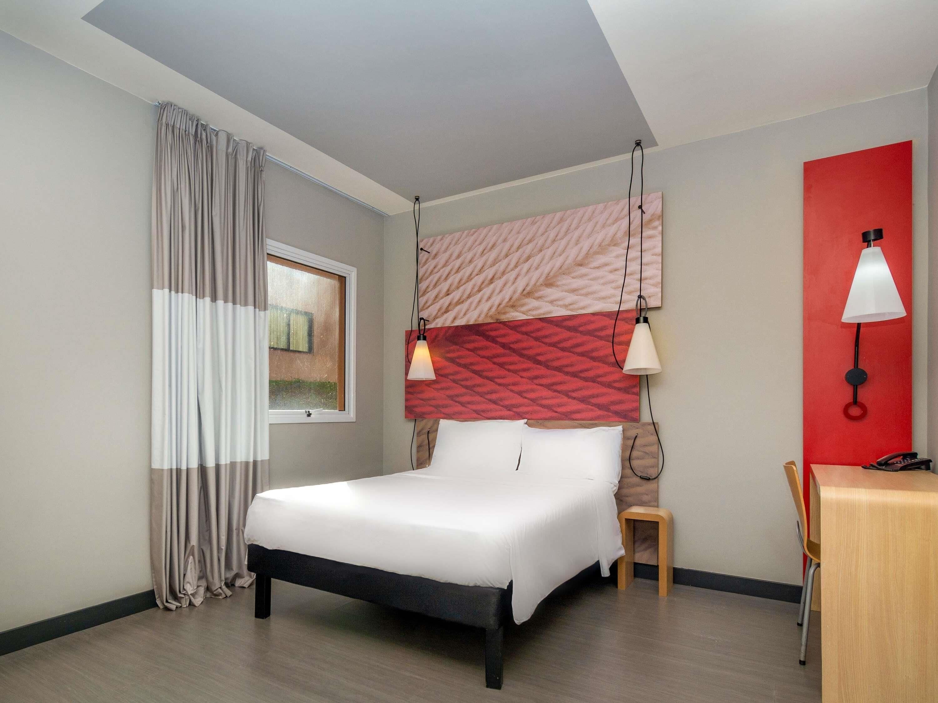 Une chambre d’hôtel comprenant un lit, un bureau, une chaise, une fenêtre avec des rideaux et un mur d’accent de couleur rouge.