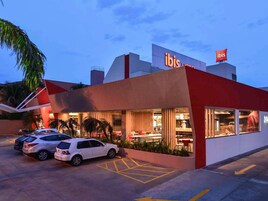 Un hotel moderno con fachada roja y blanca, un estacionamiento con coches blancos y un restaurante o zona de comedor visible con terraza.