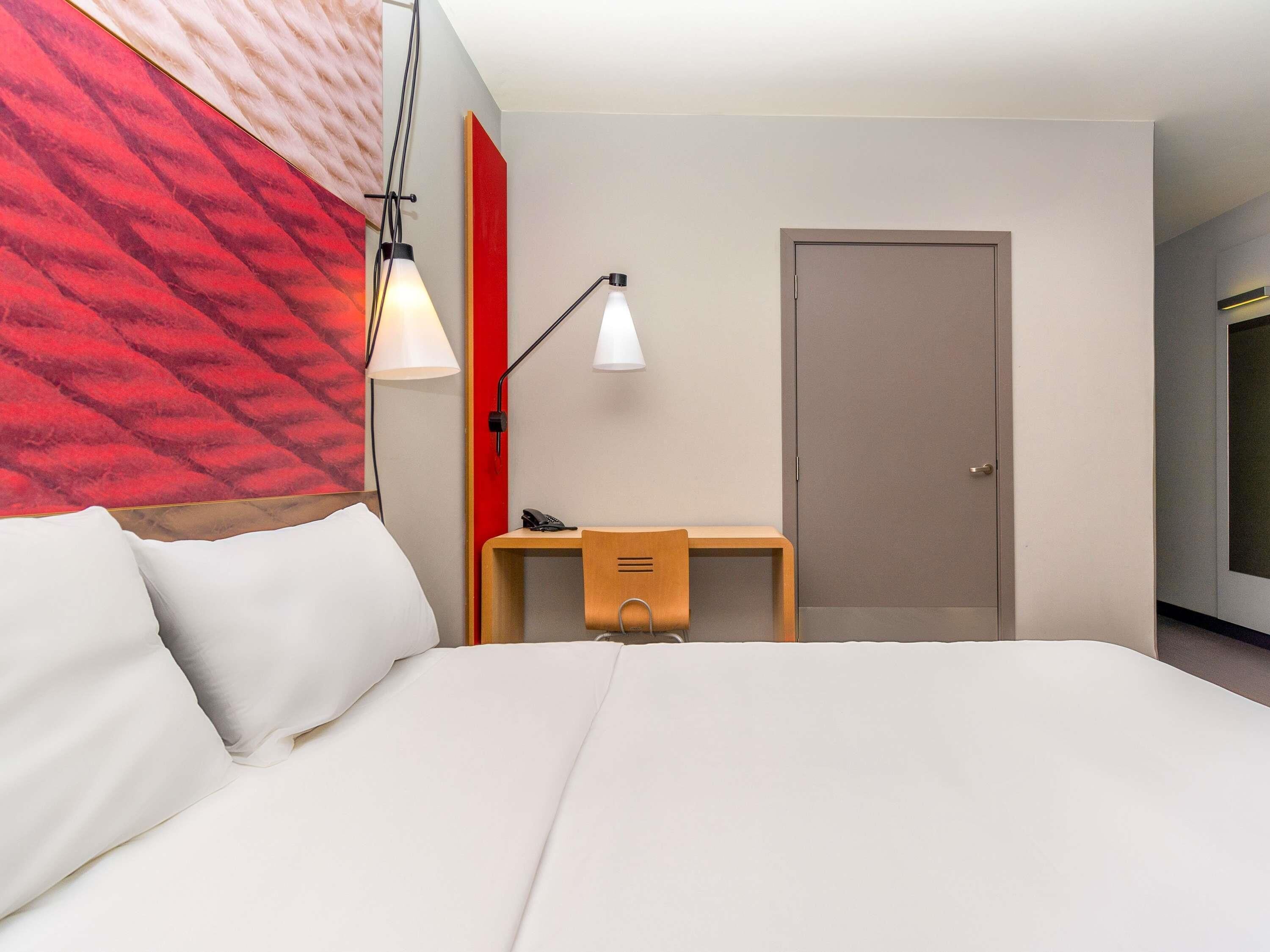 A modern hotel room with a bed, a desk, a chair, and a door.