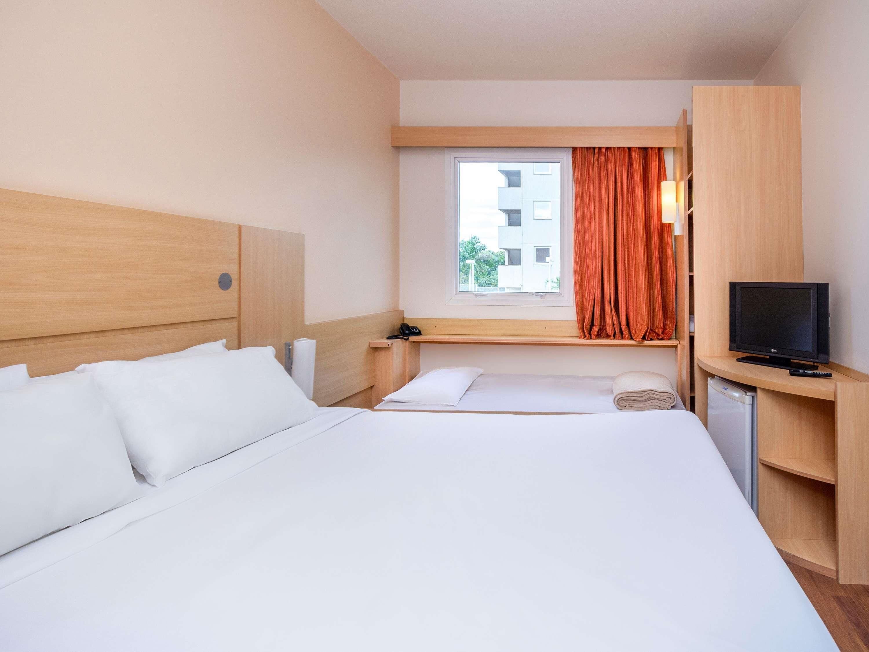 Une chambre d’hôtel avec un grand lit, une télévision, un bureau avec une lampe et une fenêtre avec des rideaux.