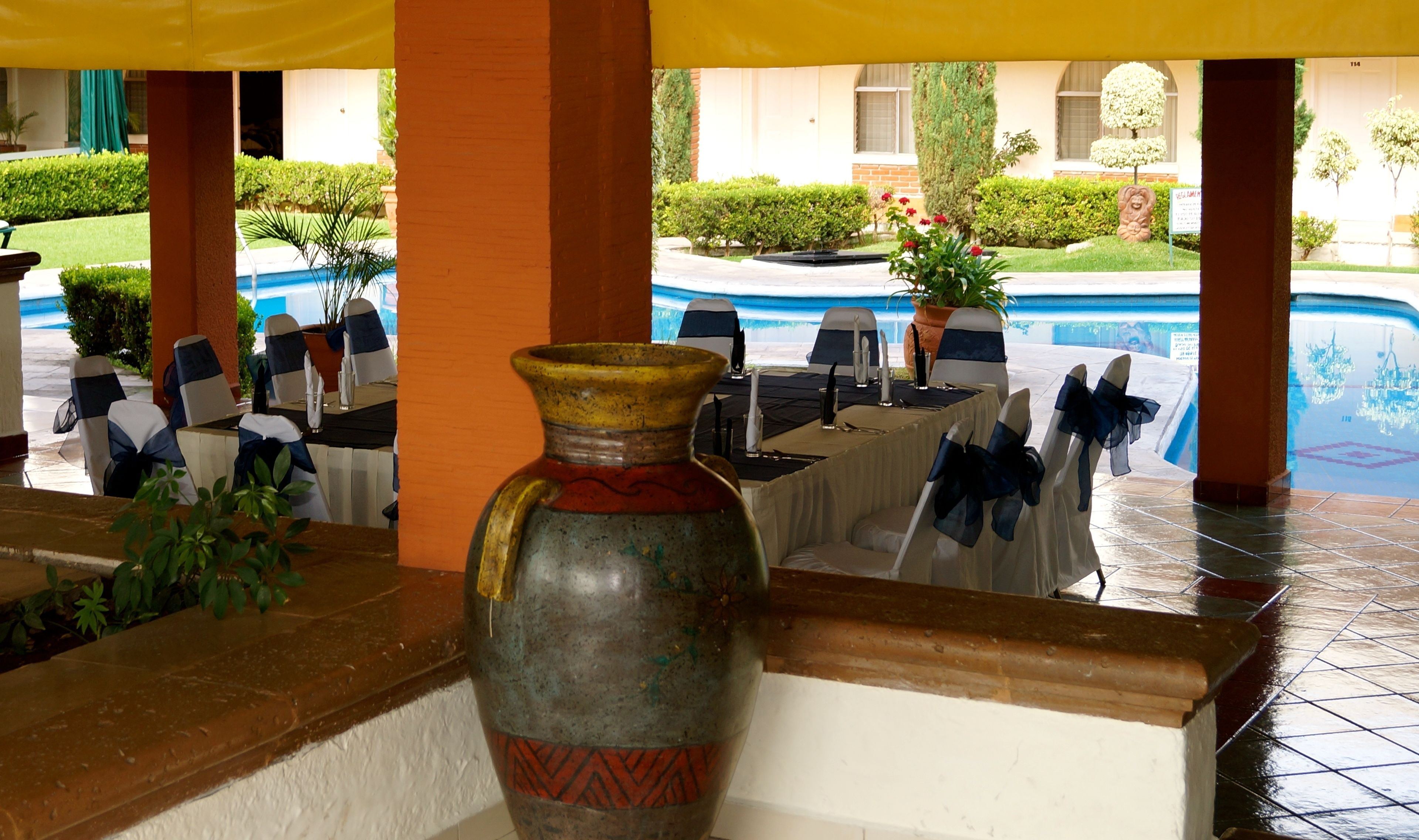 Un comedor con mesas dispuestas para una comida, un jarrón decorativo grande y una piscina al fondo.