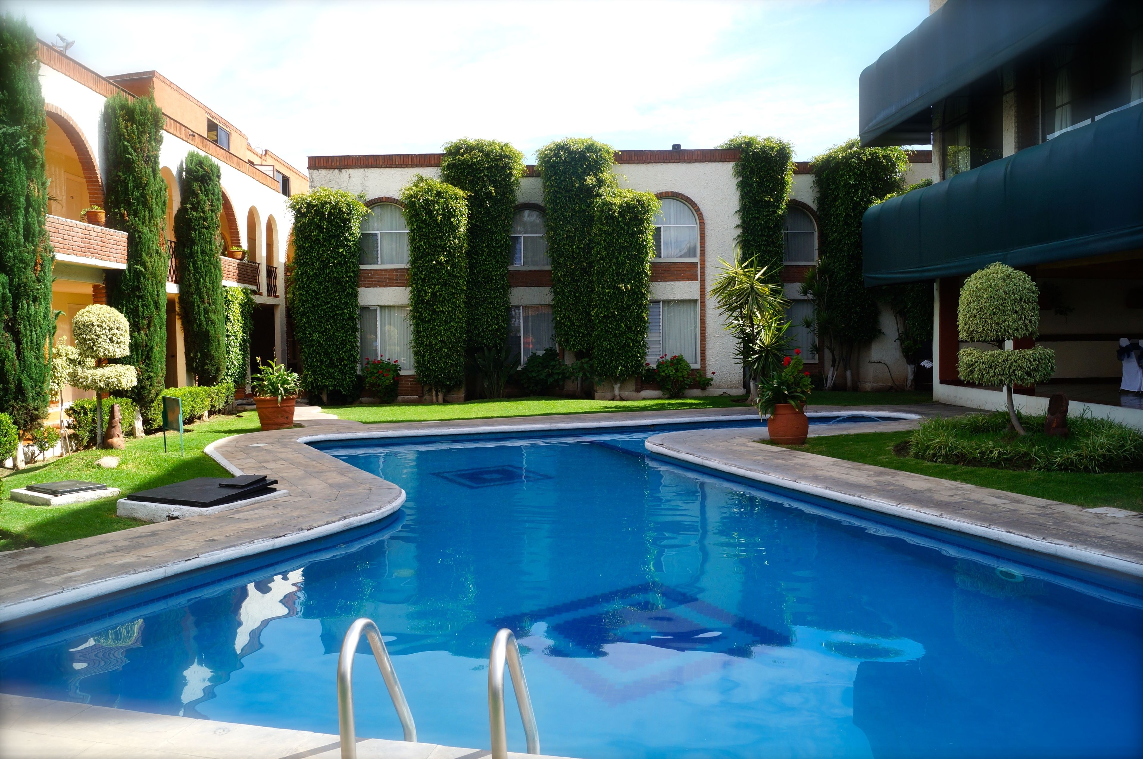 A swimming pool with a metal ladder, surrounded by well-maintained greenery and a building with multiple windows and balconies.